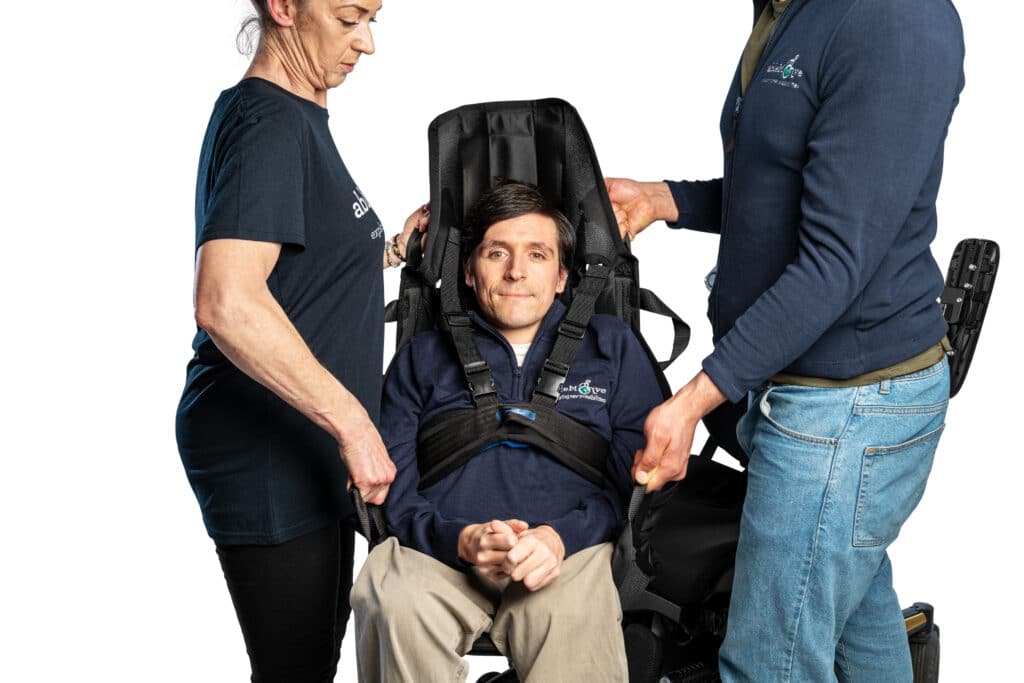 Two people are lifting a wheelchair user in a transfer sling which has a head rest. The wheelchair user is secured with a harness around their chest for extra safety