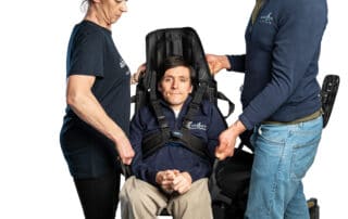 Two people are lifting a wheelchair user in a transfer sling which has a head rest. The wheelchair user is secured with a harness around their chest for extra safety