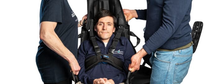 Two people are lifting a wheelchair user in a transfer sling which has a head rest. The wheelchair user is secured with a harness around their chest for extra safety